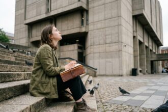 Étudiante dessinant sur un escalier en pierre face à un bâtiment universitaire brutaliste en milieu urbain